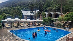 Zona de piscina con sillones de descanso y sombrillas, rodeada de edificios y árboles.
