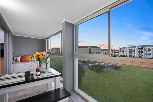 Un balcón con vistas a una zona residencial, una mesa con una botella y copas, y un florero con flores.