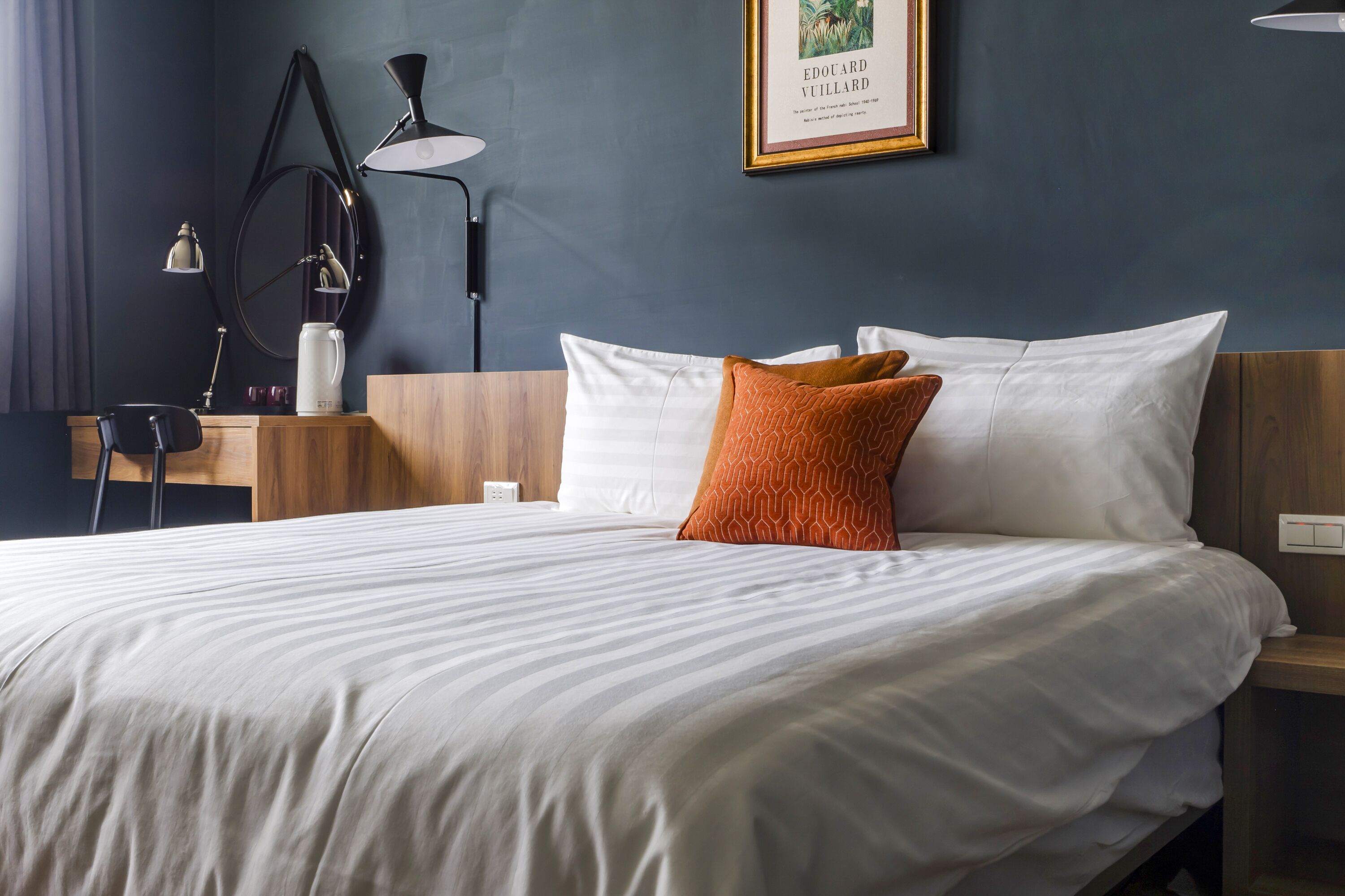 A neatly made bed with white and orange pillows, a wooden nightstand with a lamp, and a framed picture on the wall.