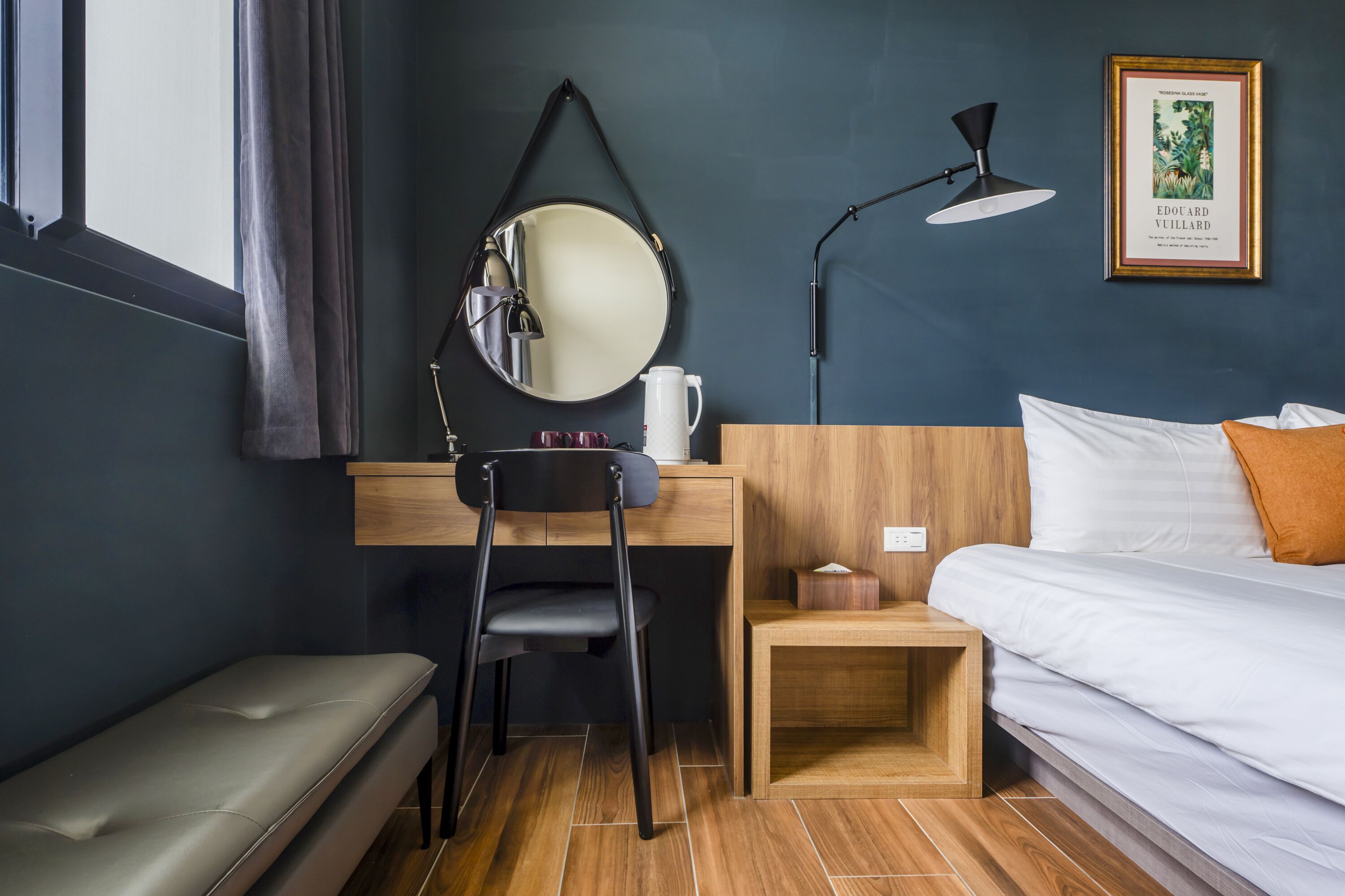 A hotel room with a bed, a desk with a chair, a small table, a mirror, a lamp, and a framed picture on the wall.