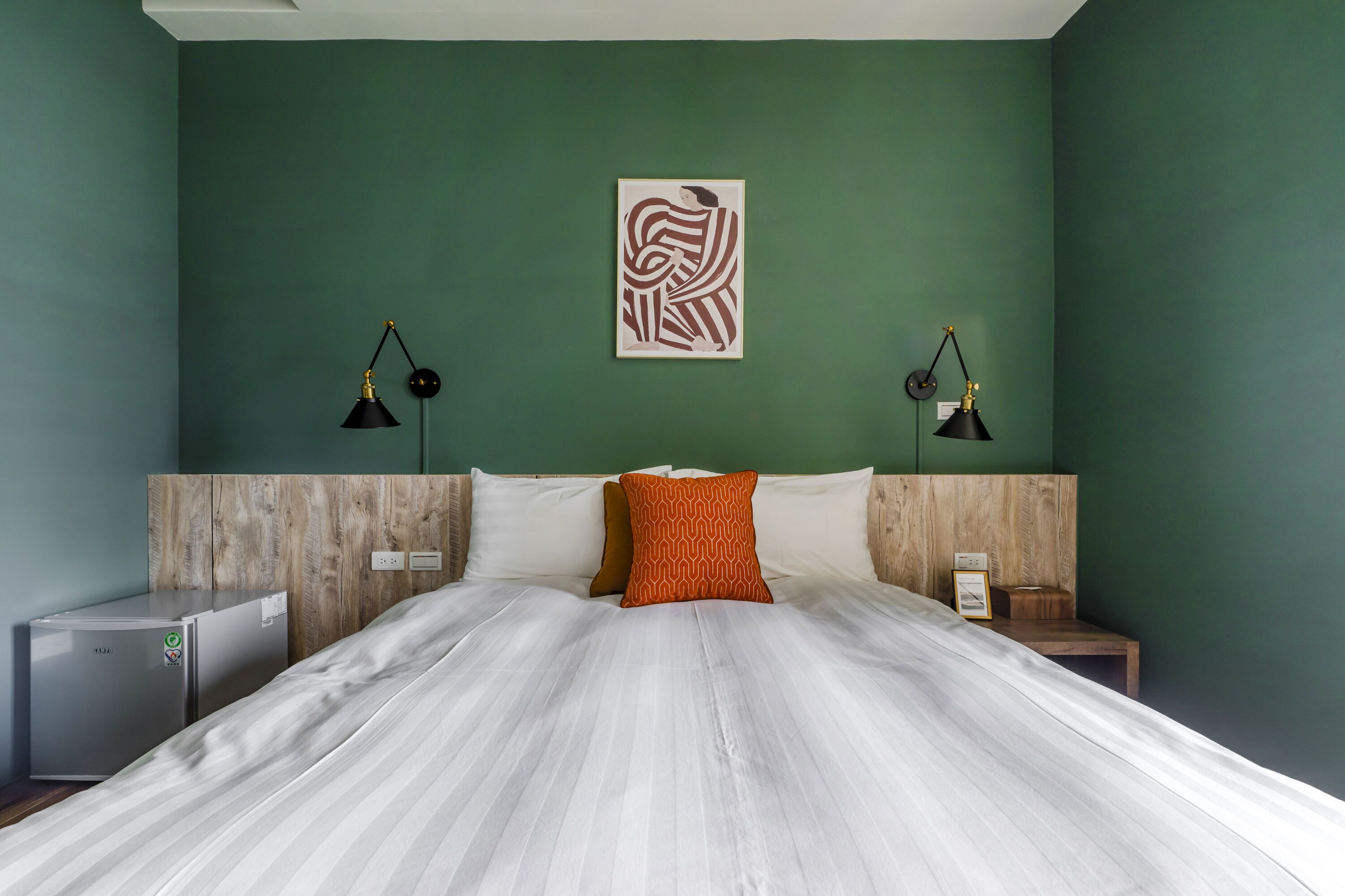 Ein ordentlich bezogenes Bett mit weißen Laken und einem orangefarbenen Kopfkissen, ein kleiner Nachttisch mit Lampe und ein an der Wand hängendes Kunstwerk.