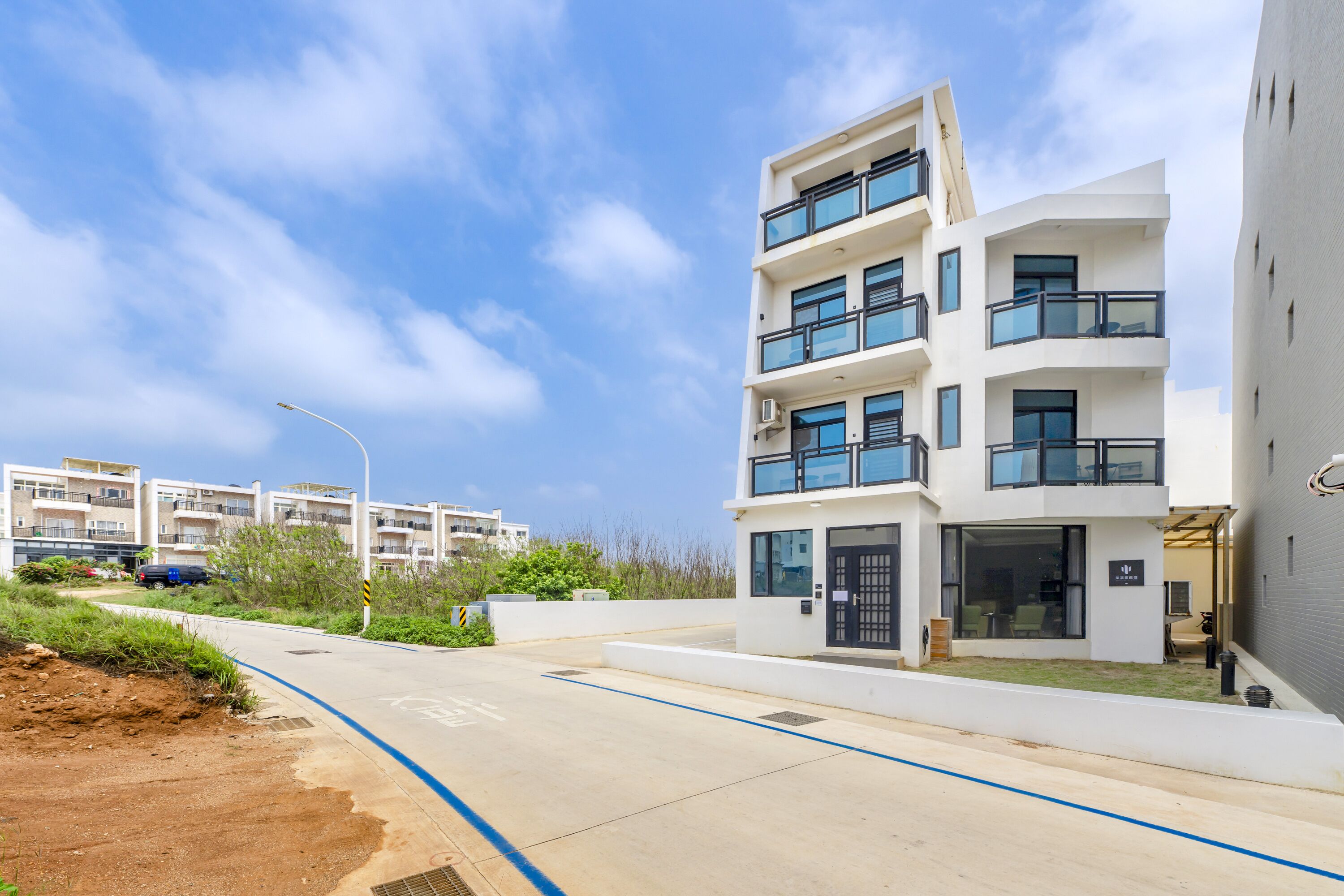 A modern multi-story residential building with balconies, a paved driveway, and a clear sky.