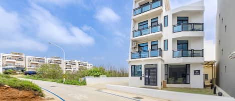 A modern multi-story residential building with balconies, a paved driveway, and a clear sky.