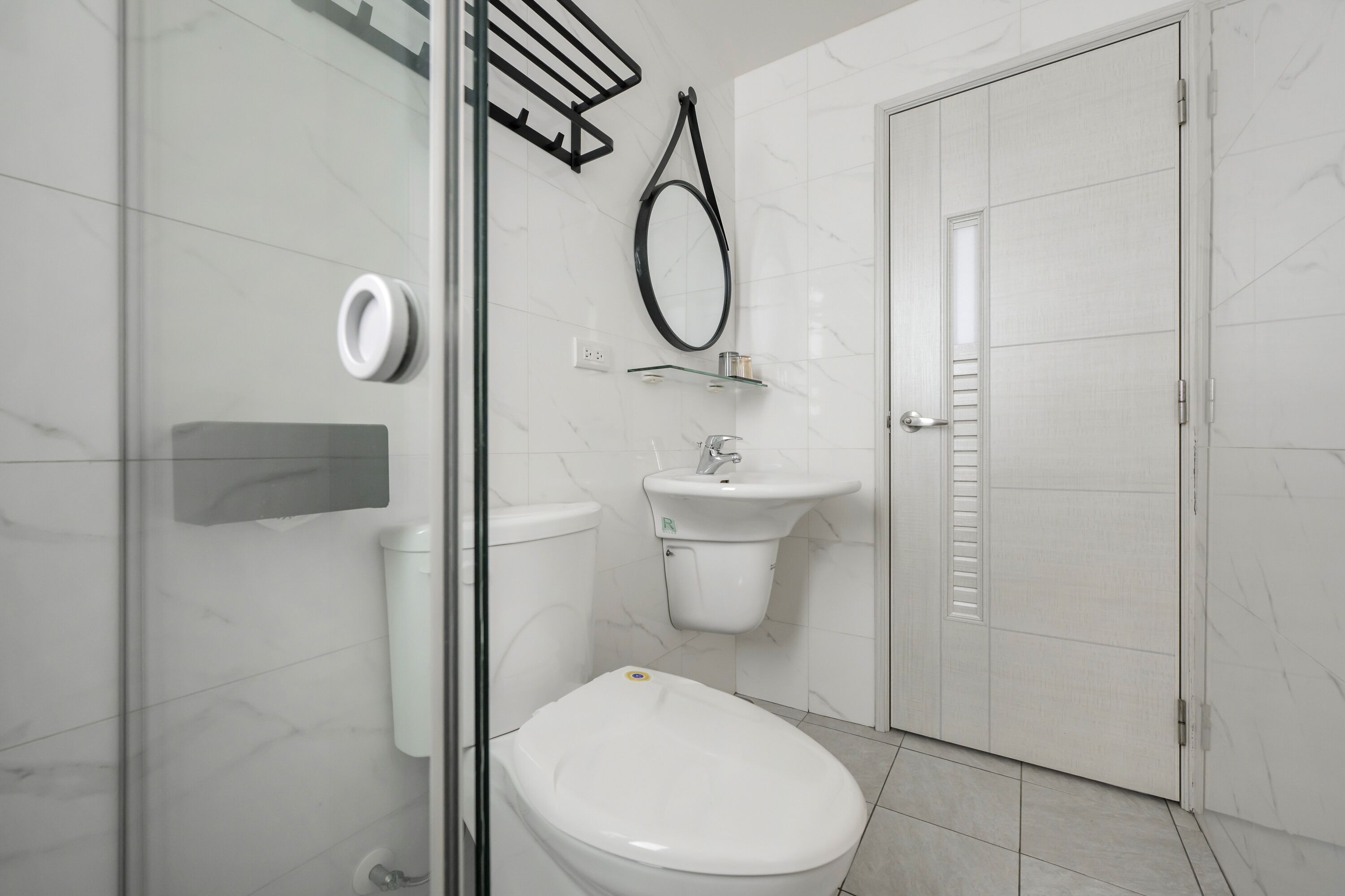 A modern bathroom with a glass shower enclosure, a white toilet, a pedestal sink, and a door with a louvered window.