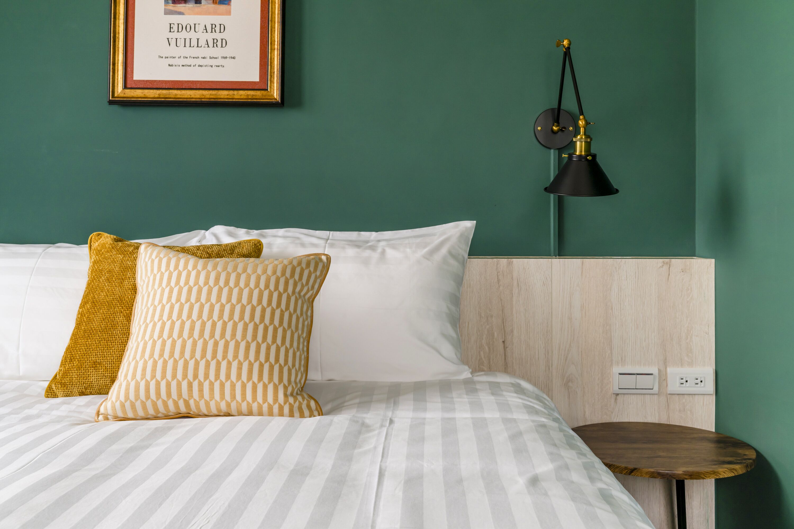 Un lit bien fait, avec une literie à rayures, deux oreillers, une petite table de chevet et une lampe fixée au mur.