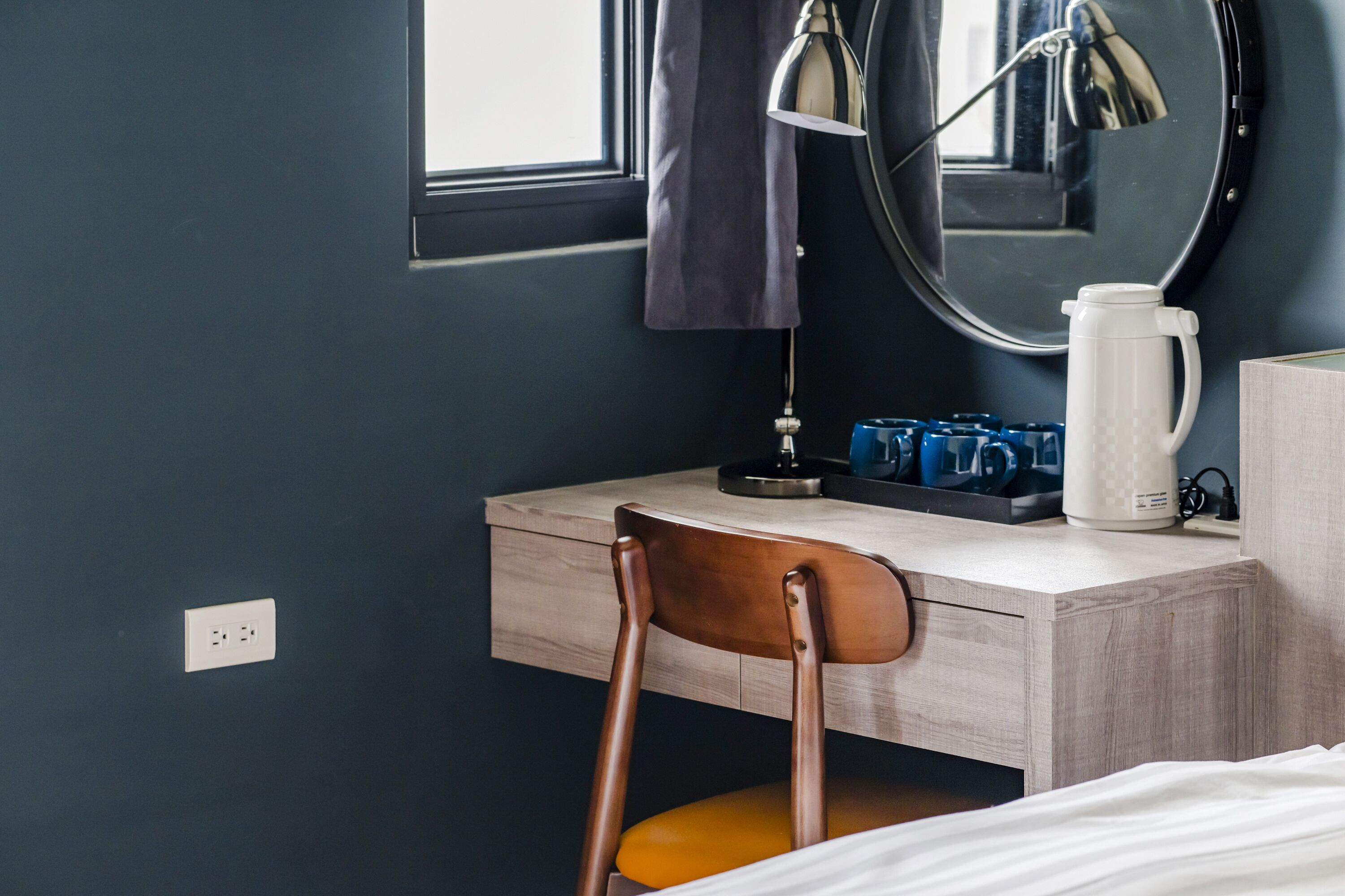 Une pièce comprenant un bureau en bois, une chaise, un miroir, une lampe, une machine à café et une fenêtre.