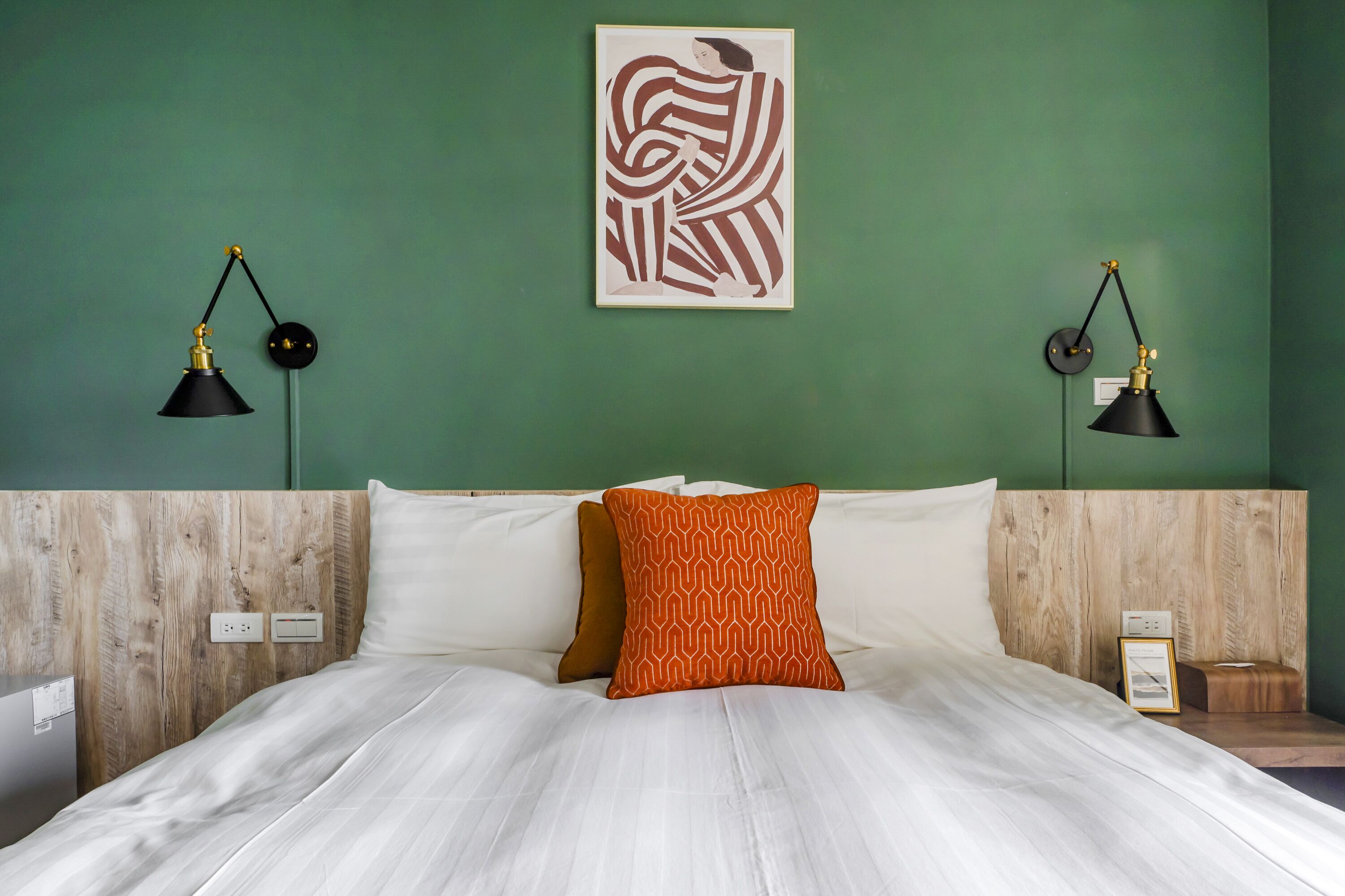 A neatly made bed with white linens and an orange patterned pillow, flanked by two black desk lamps on either side.