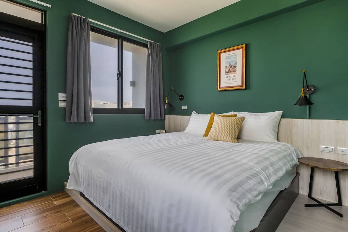 Ein Schlafzimmer mit einem großen Bett, einem Fenster mit Vorhängen, einem gerahmten Bild an der Wand und einem kleinen Tisch neben dem Bett.