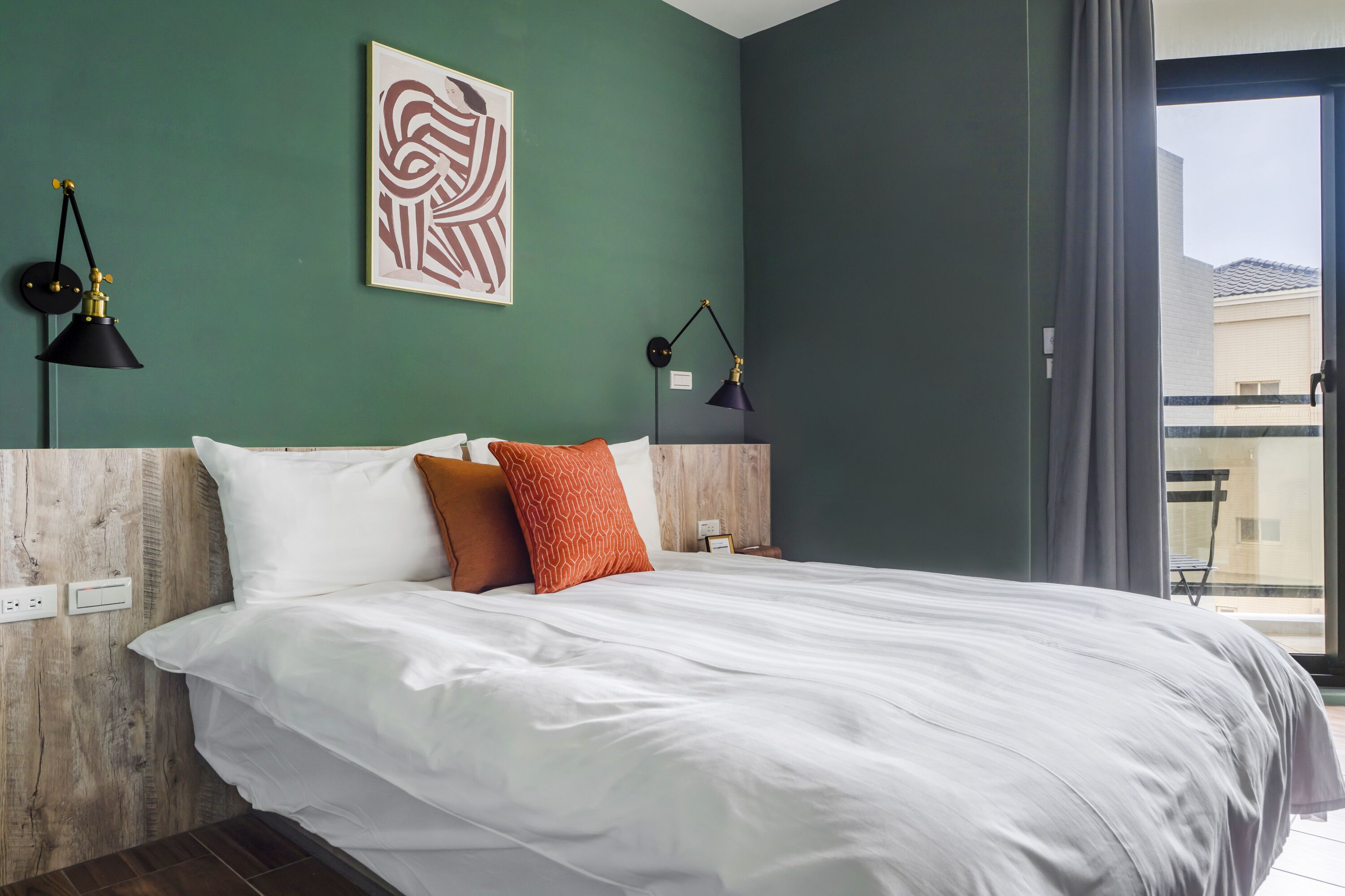 A modern hotel room with a large bed, two bedside lamps, a framed artwork on the wall, and a view of the outside through a large window.