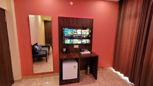 Habitación de hotel con pared roja, un espejo grande, un televisor de pantalla plana sobre un soporte de madera, un refrigerador pequeño y una puerta con marco de madera oscura.