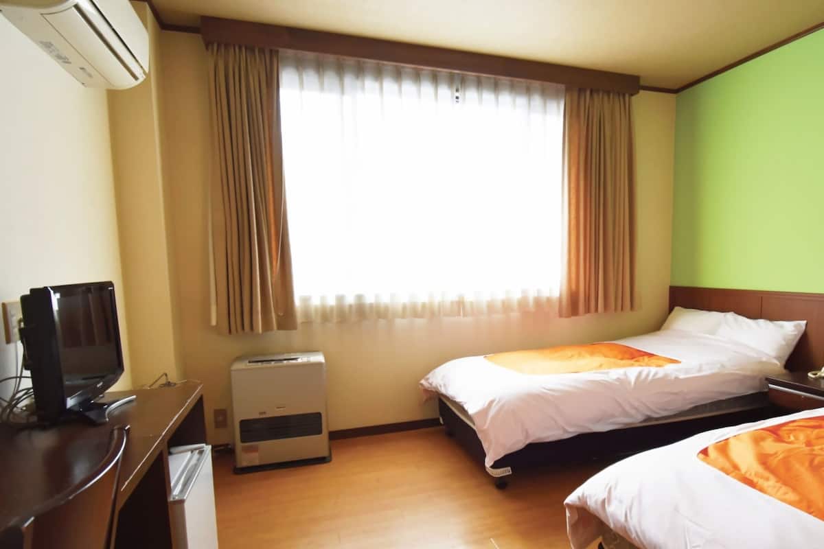 A hotel room with two beds, a desk with a computer, an air conditioner, and a window with curtains.