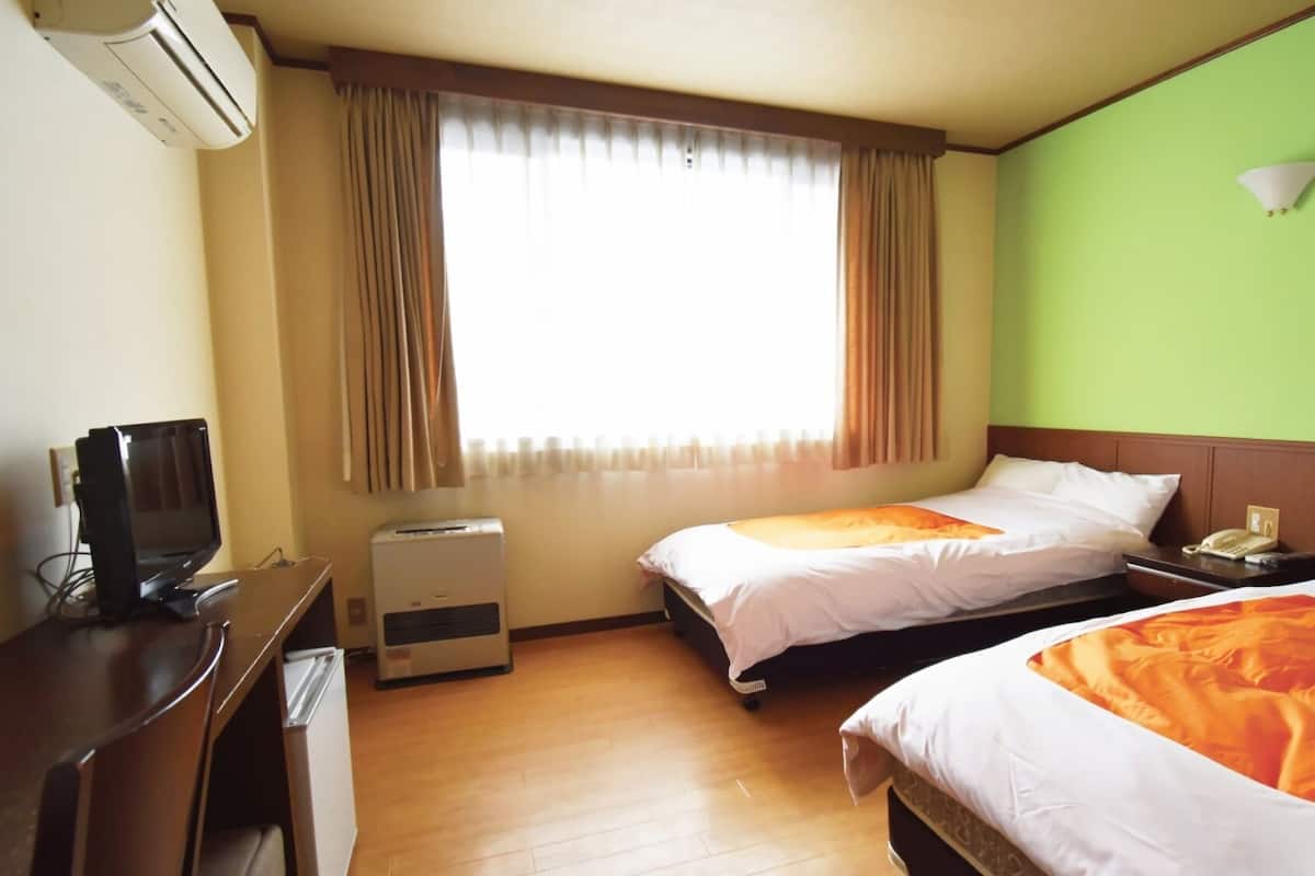 A hotel room with two beds, a desk, a chair, an air conditioner, a window with curtains, and a green wall.