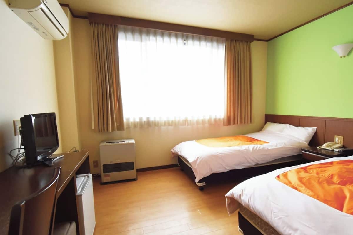 A hotel room with two beds, a desk, an air conditioner, a window with curtains, and a green wall.