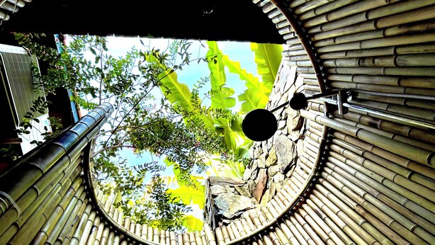 A circular bamboo structure with a central opening, a metal railing, and a view of greenery outside. - TRUONG PHU HOUSE (Hue)