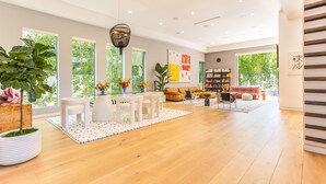 A spacious living room with a wooden floor, white furniture, and large windows offering a view of greenery outside.