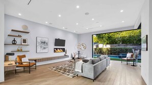 A modern living room with a grey sofa, wooden flooring, and a large window overlooking a garden. - LA Villa by AvantStay | Walk to The Grove LA (Los Angeles)