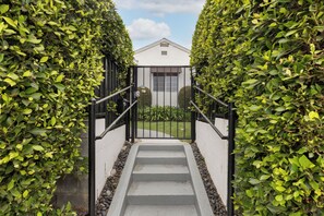 Steps lead to a modern entrance with a black gate and a small sign above, set amidst a well-manicured lawn and a small tree.