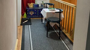A narrow hallway with a wooden ceiling, a small table with a white tablecloth, and a chair. There is a window with red curtains and a view of greenery outside.