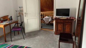 A room with a desk, chair, TV, wardrobe, and a door leading to a dining area.