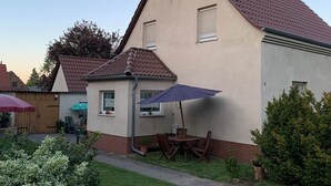A two-story house with a red-tiled roof, a small balcony, and a patio with a table and chairs.