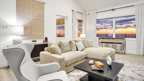 A modern living room with a large sectional sofa, a coffee table, and a view of the sunset through large windows. - Castlewood by AvantStay | Malibu Cliffside + View (Malibu)