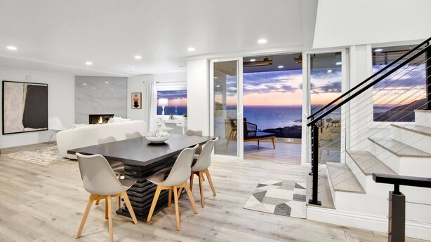 A modern living room with a dining area, a fireplace, and a large glass door leading to a balcony with a view of the ocean. - Castlewood by AvantStay | Malibu Cliffside + View (Malibu)