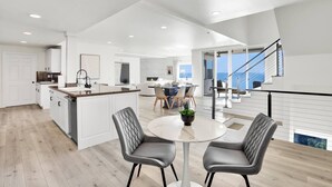 A modern kitchen with white cabinetry, a central island, and a dining area with two chairs and a round table. - Castlewood by AvantStay | Malibu Cliffside + View (Malibu)