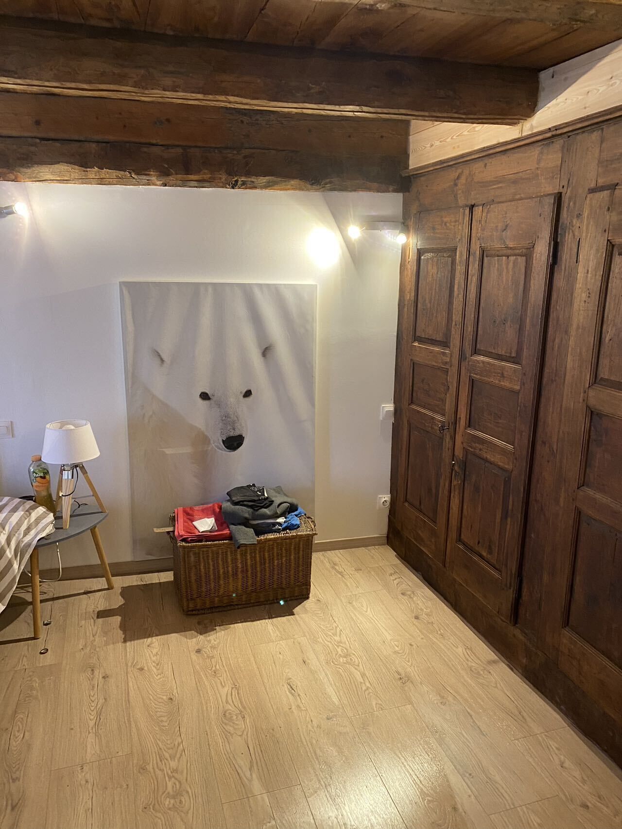 Une pièce avec un plancher en bois, un lit recouvert d’une couverture à rayures, une petite table avec une lampe, un panier rempli de vêtements et une armoire en bois.