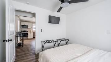 A bedroom with a bed, a ceiling fan, a TV, and a kitchenette in the background.