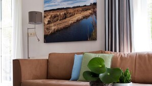 A living room with a brown leather sofa, a wall-mounted lamp, a framed landscape painting, and a potted plant on a white table. - Bungalow near De Alde Feanen Reserve (Earnewald)