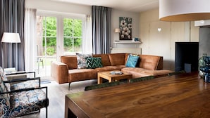 A modern living room with a sofa, coffee table, and a dining table. There is a window with curtains, a lamp, and a painting on the wall.