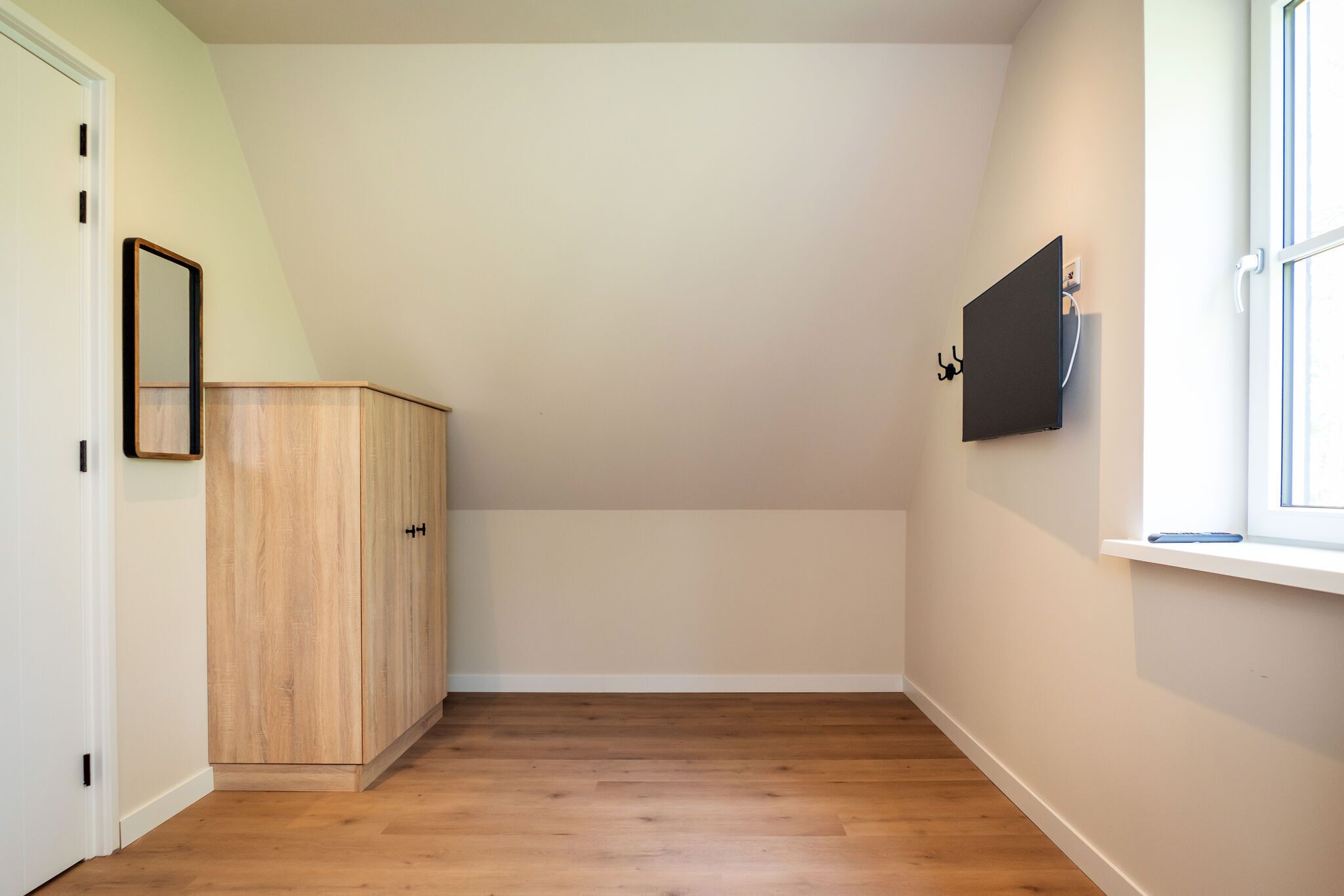 A room with a wooden floor, a wardrobe, a TV mounted on the wall, and a window.
