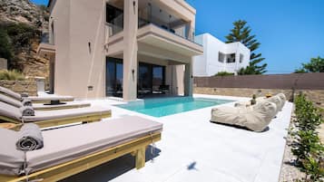 A modern two-story house with a swimming pool, sun loungers, and a stone wall.