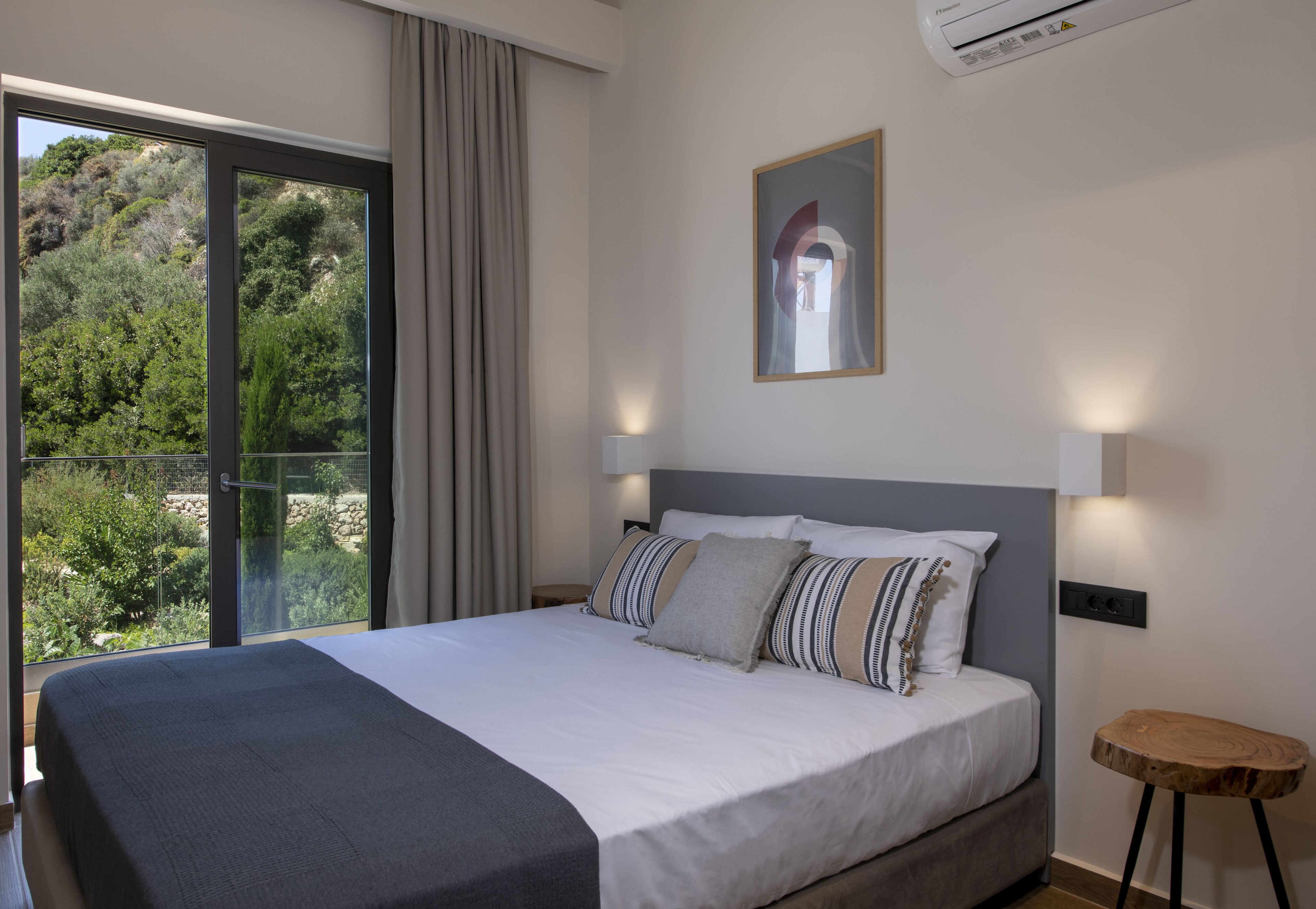 A bedroom with a large bed, a view of greenery through a sliding door, a framed picture on the wall, and a small wooden stool.