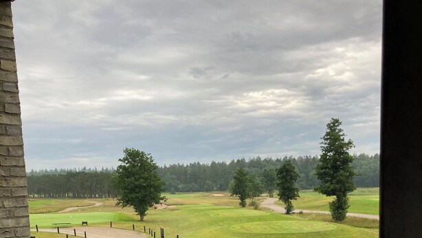 Vista desde un balcón sobre un campo de golf, con un muro de ladrillo, barandilla metálica y suelo mojado.
