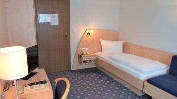 Ett hotellrum med en säng, ett skrivbord med en lampa, en stol, ett litet bord och en telefon som är monterad på väggen.