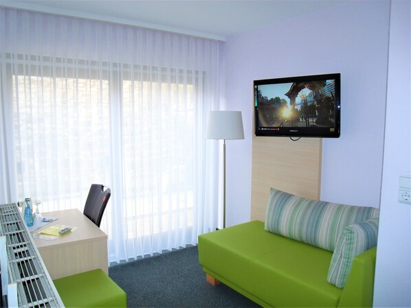 Habitación de hotel con sofá verde, escritorio y una silla, televisor montado en la pared y ventana con cortinas translúcidas.