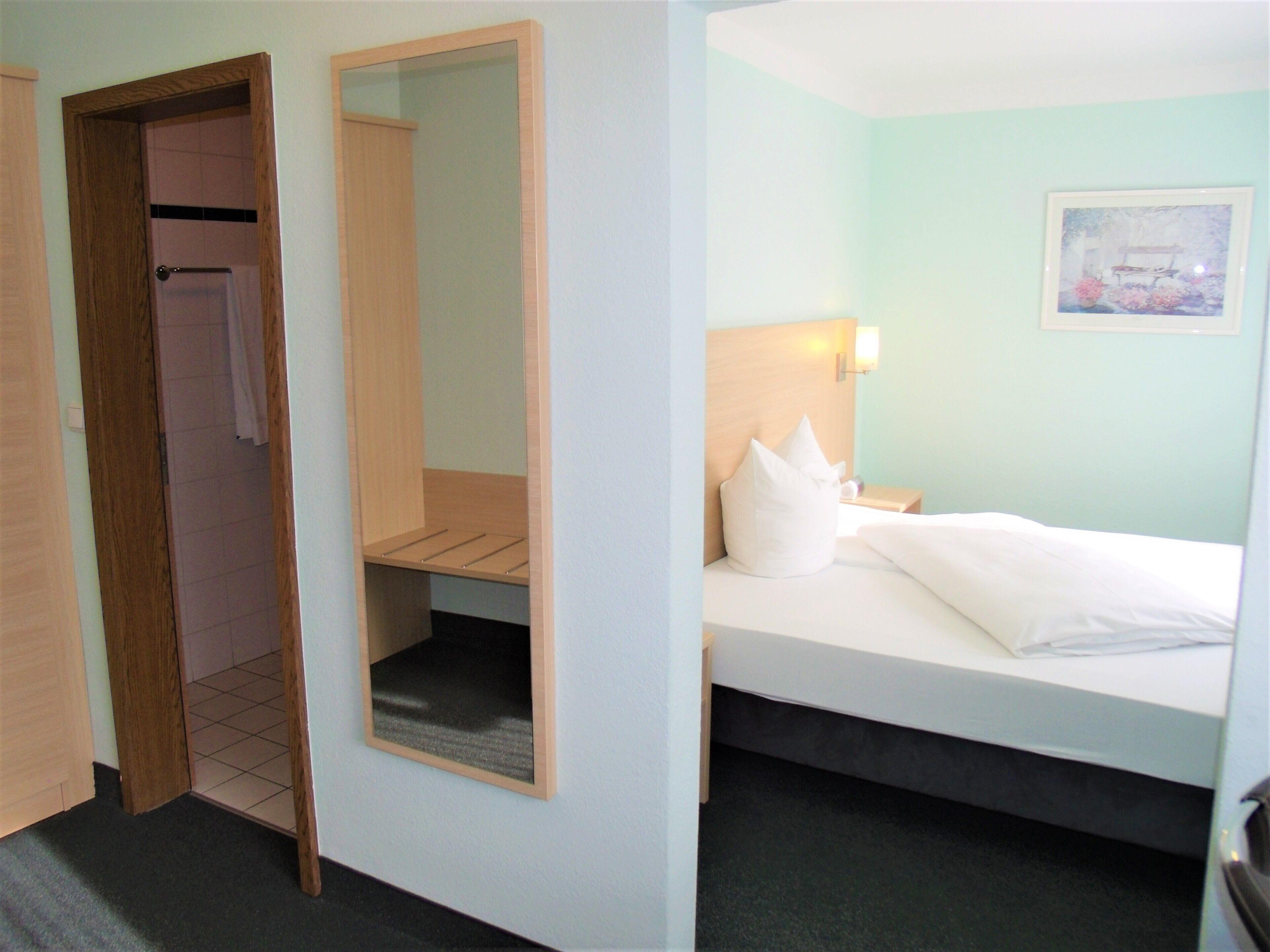 A hotel room with a bed, a nightstand, a wall-mounted lamp, a framed picture, a wooden wardrobe, and a mirror.