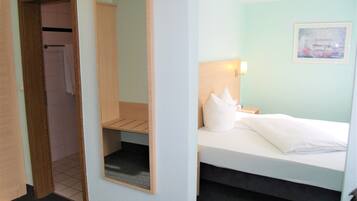 A hotel room with a bed, a nightstand, a wall-mounted lamp, a framed picture, a wooden wardrobe, and a mirror.