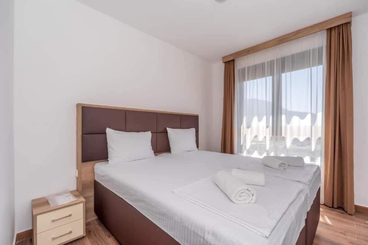 Cama bem arrumada com lençóis brancos e uma toalha enrolada, mesa de cabeceira de madeira com gavetas, e uma janela grande com cortinas leves.
