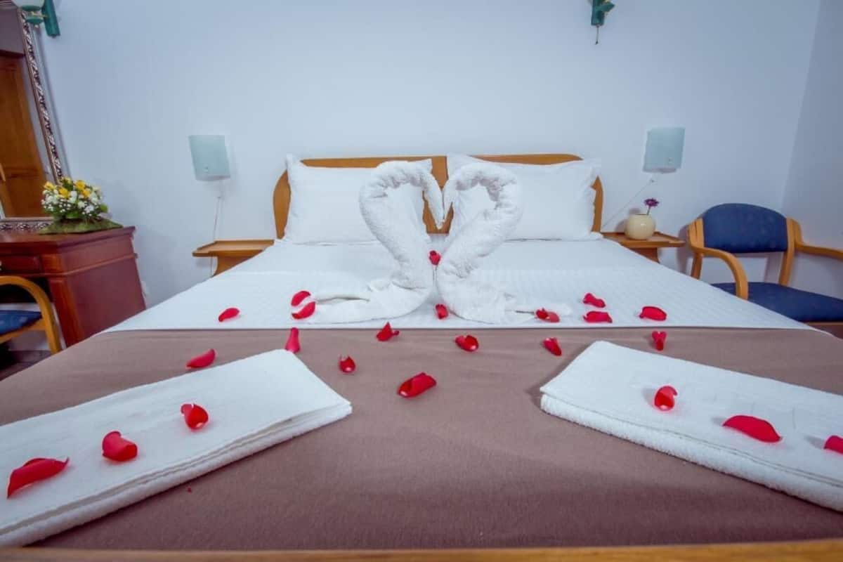 Quarto de hotel com cama bem arrumada, cabeceira de madeira, mesa de cabeceira com abajur, uma cadeira e cômoda com espelho e flores.
