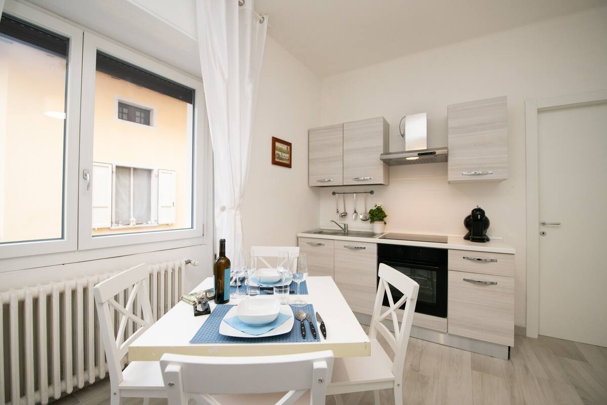 Una cocina moderna con armarios blancos, una mesa de comedor preparada para cuatro personas y una ventana con cortinas blancas.