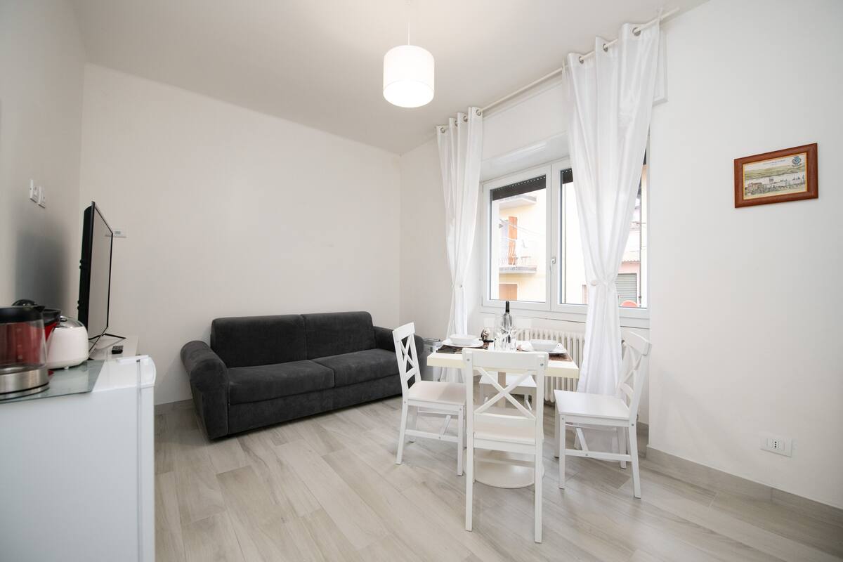 Una sala de estar moderna con un sofá gris, una mesa de comedor blanca con sillas, un radiador blanco y una ventana con cortinas blancas.