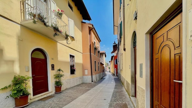 A narrow street with buildings on either side, featuring a red door and a wooden door on the right. - La Torretta (Moniga del Garda)