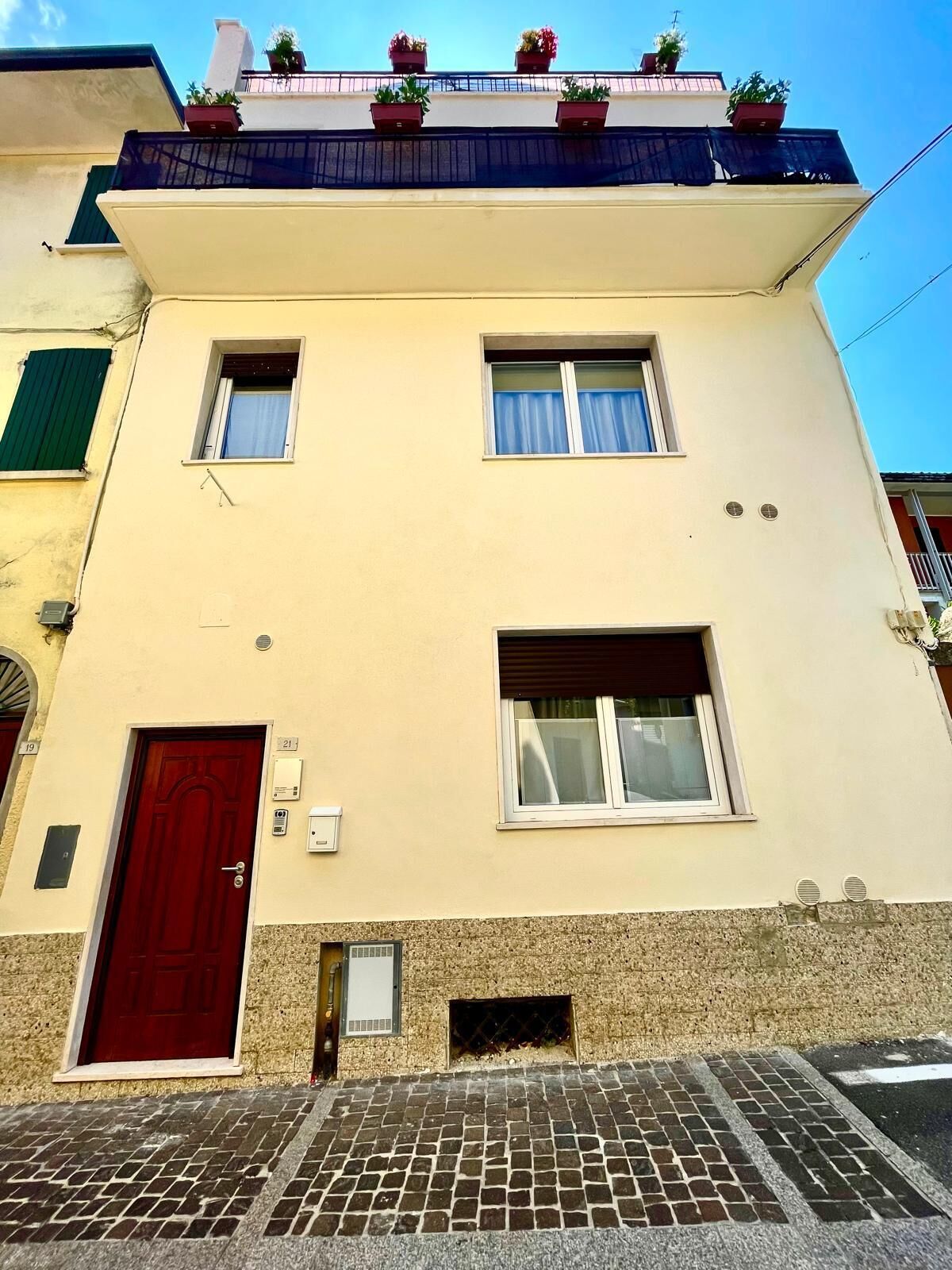 Un immeuble de deux étages avec une porte rouge, portant le numéro 21, et une petite fenêtre avec un rideau.