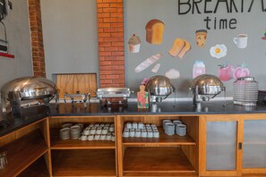 A breakfast bar with a brick wall and wall art, buffet setup with food and drink containers, and wooden shelves with cups.