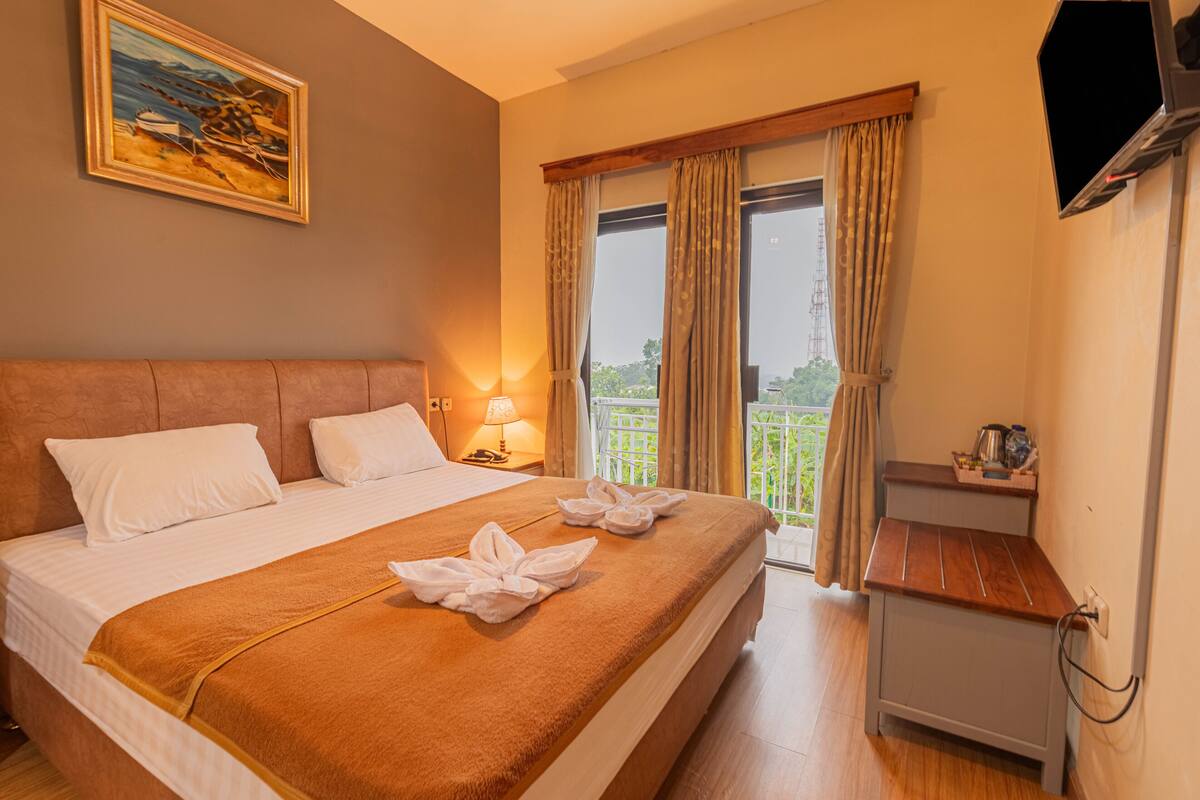 Ein Hotelzimmer mit einem großen Bett, einem Gemälde an der Wand, einem Balkon mit Vorhängen und einem kleinen Tisch mit einer Lampe.