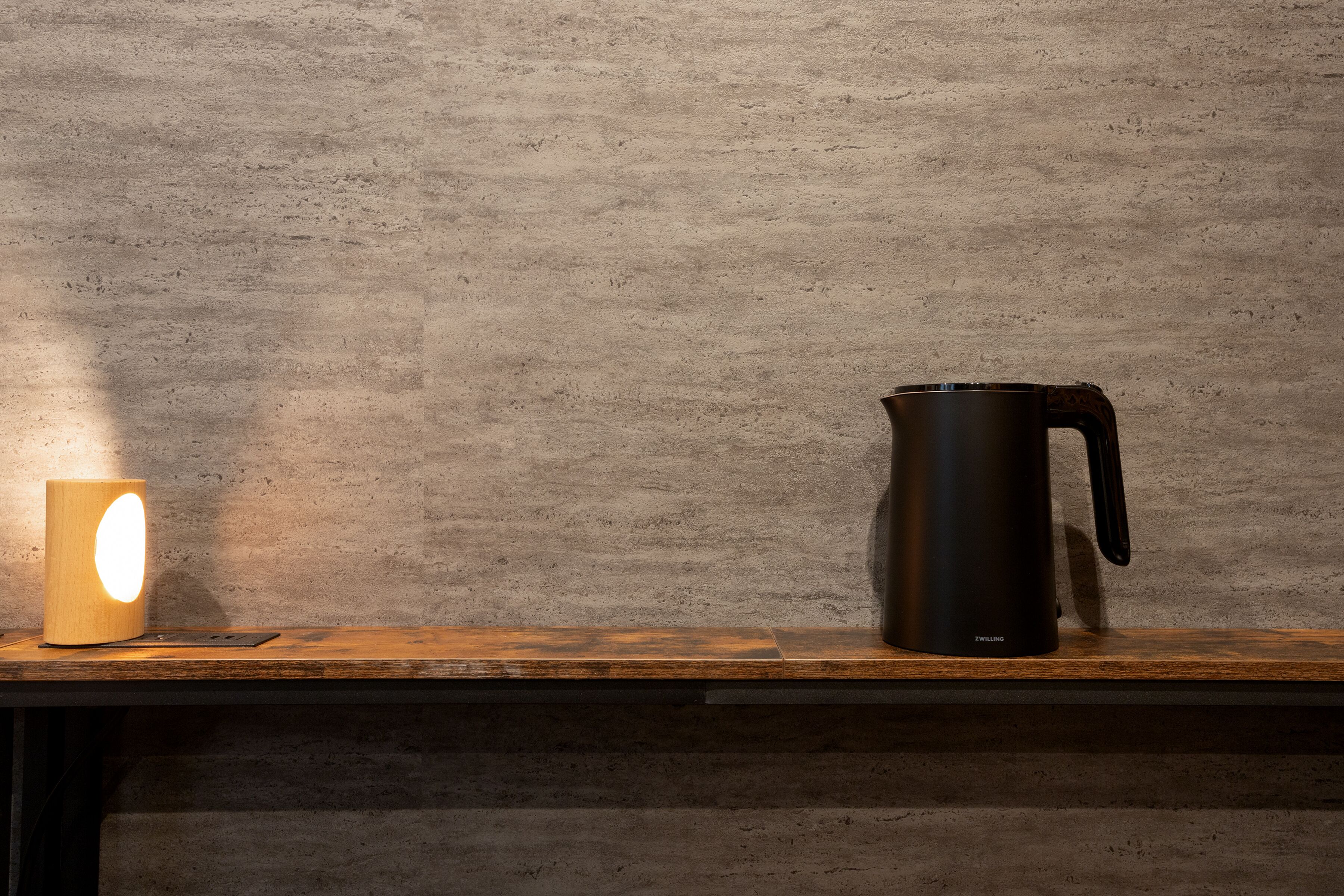 A wooden shelf with a black electric kettle and a cylindrical light source.