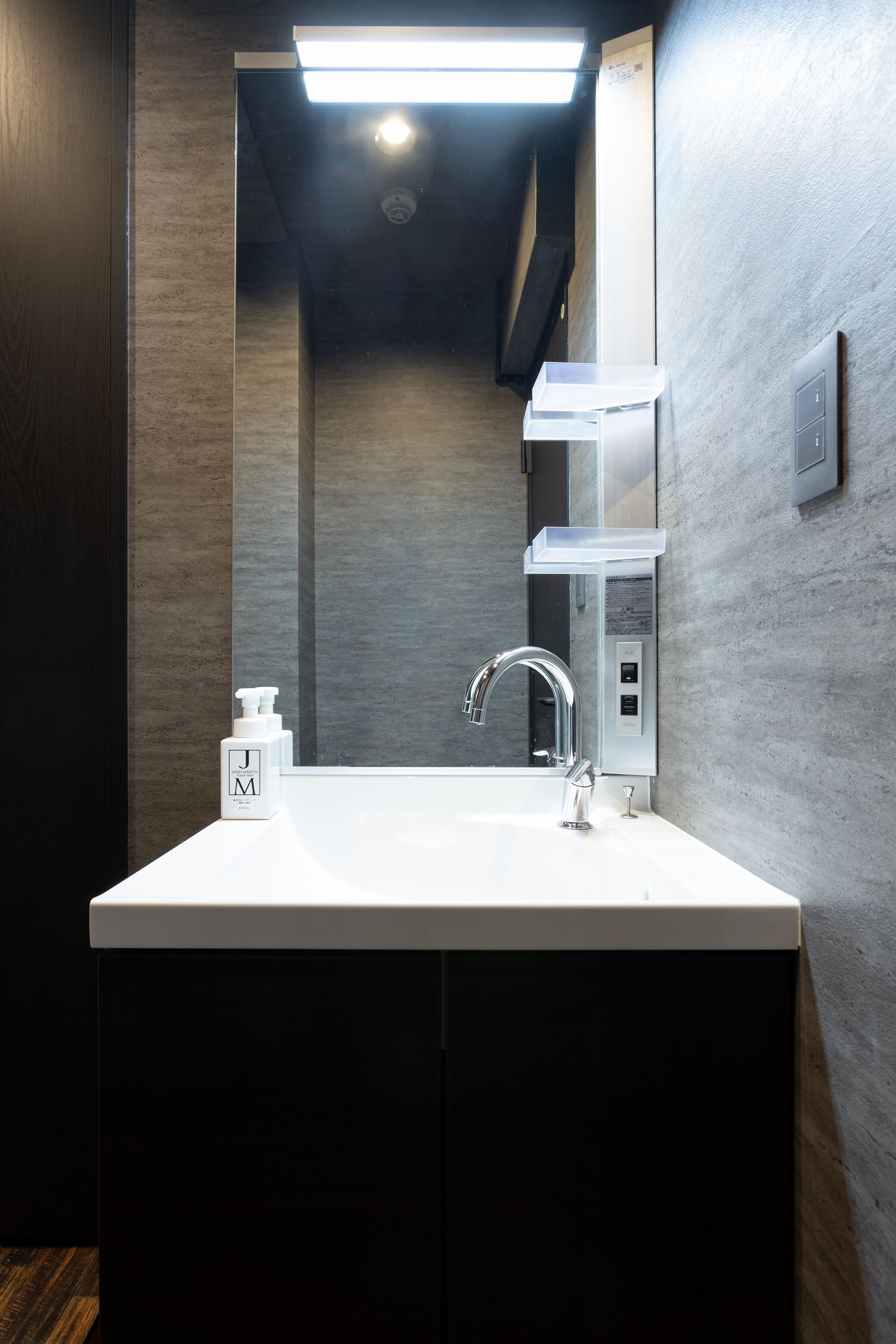 Une salle de bain moderne avec un lavabo blanc, un robinet chromé et un miroir au-dessus. Un distributeur de savon se trouve à gauche et un porte-serviettes à droite. Les murs sont texturés et un luminaire est placé au-dessus du miroir.