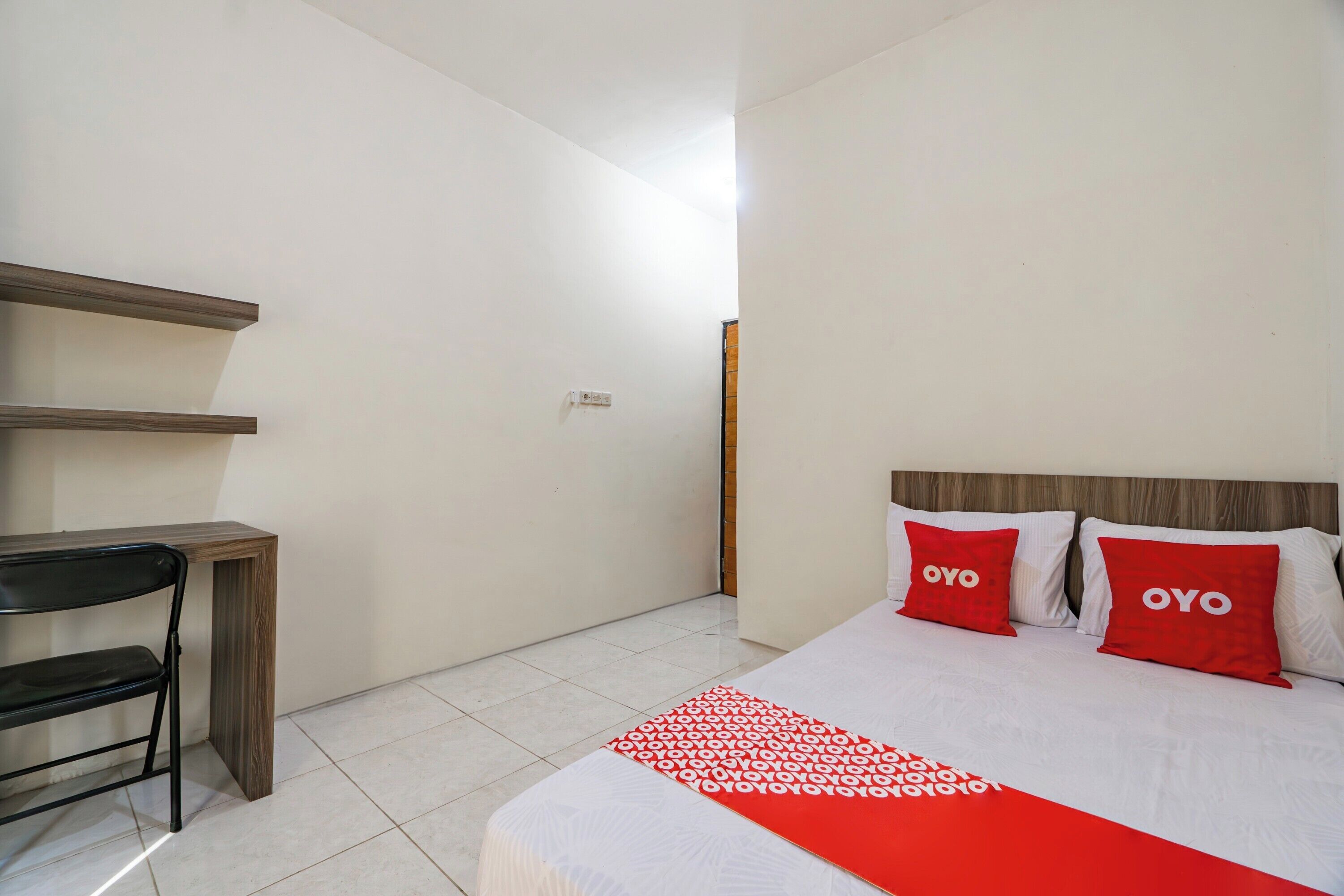 Ein Hotelzimmer mit einem Bett, zwei rote Kissen mit dem Schriftzug "OYO", einem weißen Bettüberwurf, einer roten und weißen Decke, einem Holztisch und einem schwarzen Stuhl.